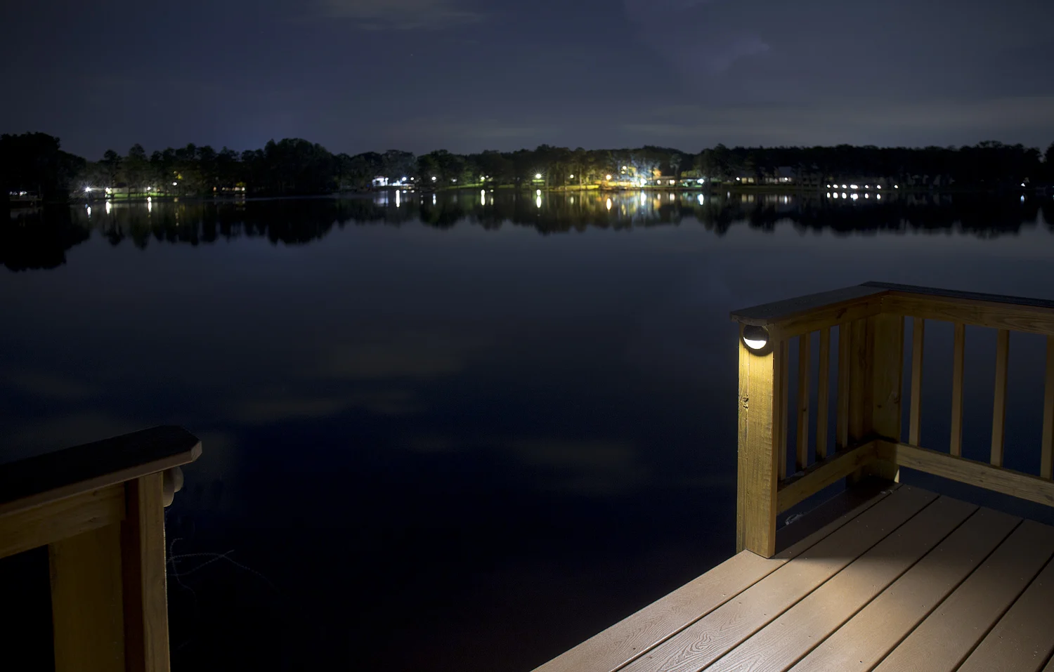 Warm post lights illuminate a wooden deck overlooking a lake at night.